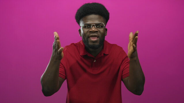 Man wearing glasses and red polo shirt gestures with both hands toward camera in a pink studio; frustration urgency.