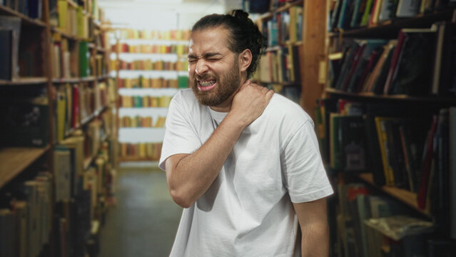 Man holding his neck with one hand among library shelves, grimacing with closed eyes and braced shoulder; pain.