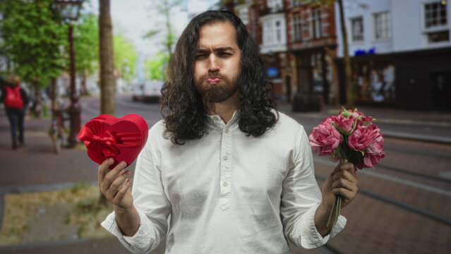 Man holds red heart box and pink bouquet in hands while puffing cheeks on a busy city street; awkward romance.