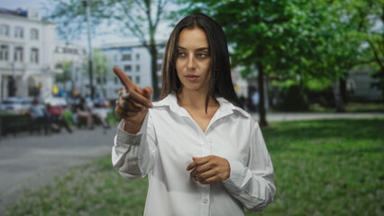Fototapeta premium Woman points finger forward on city street near building, white shirt visible and fingertip aimed at camera; determination.