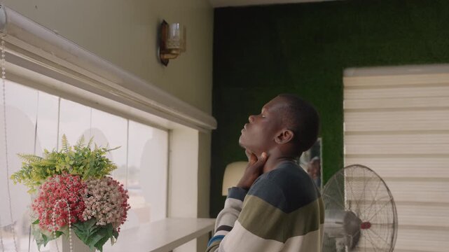 Black man touching face at window, nervous anticipation, floral bouquet on windowsill, striped sweater, warm lamp glow, soft blinds, oscillating fan, rehearsing lines as aspiring poet, hopeful gaze,