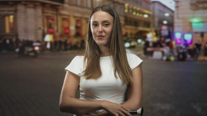 Fototapeta premium Woman holding a makeup brush with folded arms on a busy city street outside illuminated storefronts at dusk, slightly smiling; confidence.