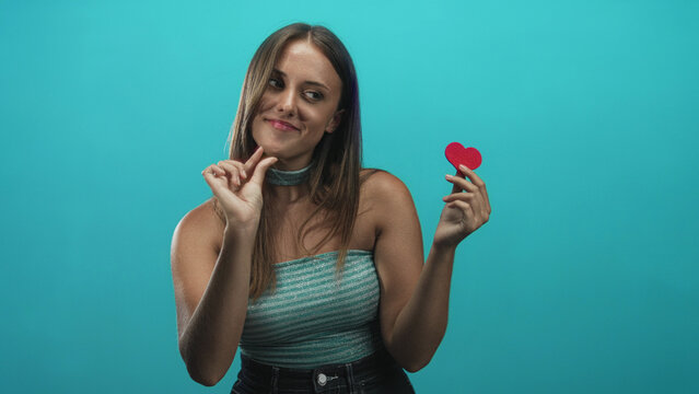 Woman holding a red heart and pinching fingers to show small size while wearing a turquoise strapless top in a studio; playful flirtation.