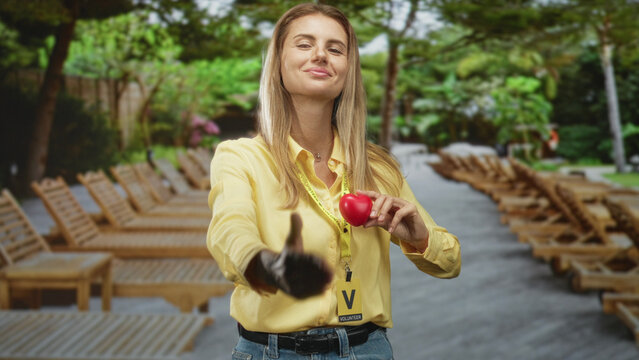 Blonde woman extends hand for handshake holding a red heart and volunteer badge at a resort pool deck; outreach caring friendly.