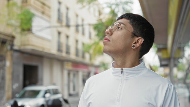 Man wearing glasses and white zip jacket looking up on street near storefront and building facade; curiosity.