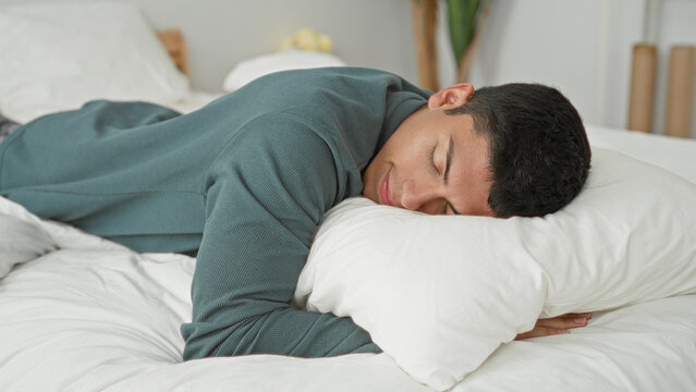 Man lying prone on bed, sleeping face on a white pillow with arm tucked under pillow in a studio bedroom set wearing a green sweater; calm rest.