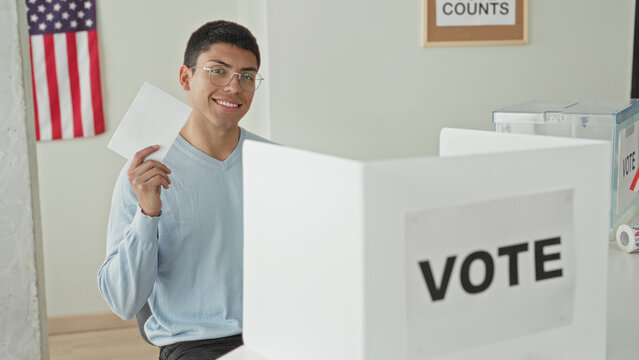Man holding ballot in hand at voting booth inside building, smiling and raising paper; civic duty pride.