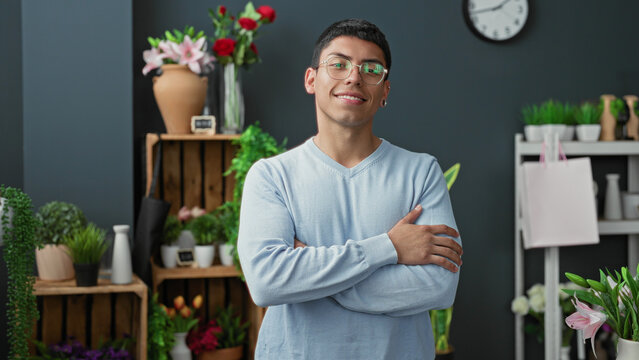 Man florist smiling with arms crossed, hand on upper arm among potted plants, vases and roses in a studio; warm confidence.