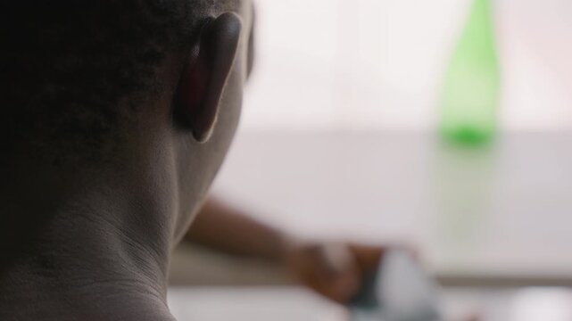 Black man checking phone screen from rear profile closeup, neck and ear in shallow focus with bokeh bottle on table, finger tapping to reply, quiet introspective mood suggesting waiting for romantic
