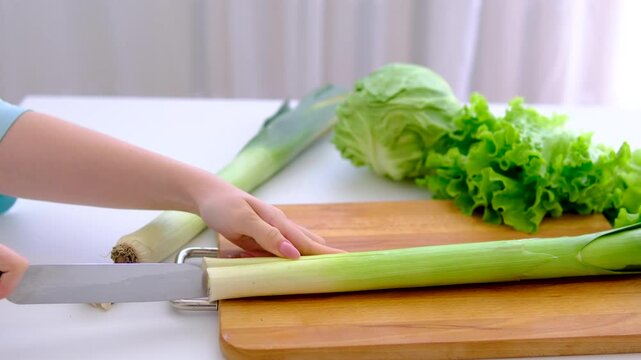 Slicing fresh leek on wooden board in bright white kitchen with cabbage and lettuce