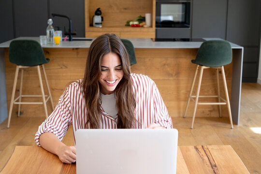 Smiling woman working on a laptop at a kitchen table, remote work from home, casual striped shirt, bright modern kitchen, video call or studying, relaxed productive lifestyle