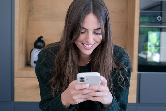 Young woman smiling and texting on smartphone in modern kitchen, relaxed at home, casual lifestyle moment with long hair, cozy sweater and natural light