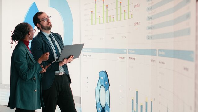 Corporate managers evaluate fiscal performance metrics on digital wall, collaborating, Diverse executive team discusses quarterly earnings report and shareholder equity charts during boardroom.