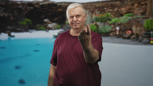 Man showing middle finger with bare hand at poolside near building, wearing maroon shirt and serious expression; defiance.