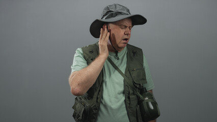Obraz premium Man fisherman wearing fishing vest and wide hat touches ear with hand in studio while holding canteen strap and looking aside; listening concentration.