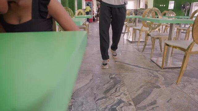 Woman seated at green table waits while partner walks toward bistro, lowangle closeup of legs and shoes on marble floor casual black outfit, row of chairs and table, cinematic anticipation