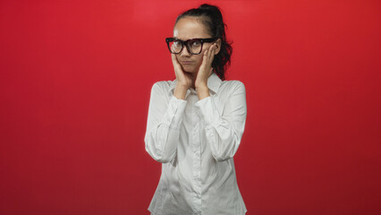 Fototapeta premium Woman wearing oversized glasses touches her cheeks and lifts her hands in a red studio setting, adjusting frames and shrugging in subtle poses; uncertainty reflection.