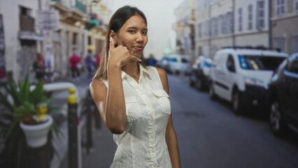Fototapeta premium Thai woman shows call me gesture with thumb and pinky at ear, bare hand visible in a white sleeveless shirt on a street; playful charm.