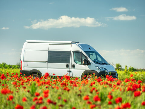 view to white camper van in red poppies field under sky with light clouds in summer day