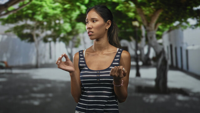 Thai woman in a striped tank top showing left hand forming ok sign and clenched right fist on a tree lined street near a building wall; skepticism reflection.