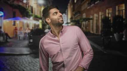 Obraz premium Hispanic man with beard looking up, hand in pocket, pink button shirt on cobblestone street with outdoor cafe lights; quiet wonder.