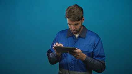 Hispanic man in blue work overalls taps tablet screen while holding device and looking down in studio  focus productivity. © Krakenimages.com