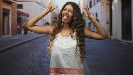 Fototapeta premium Hispanic teenage girl smiling, shows rock horns with both hands, wearing a white embroidered sundress and braces, on a cobblestone street lined with buildings; playful moment.