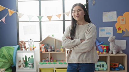 Young chinese woman with arms crossed beside a toy shelf and stuffed animals in a preschool...