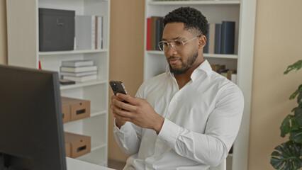 Fototapeta premium Man holding smartphone, fingers tapping screen while seated at desk with computer monitor visible in building; focused productivity.