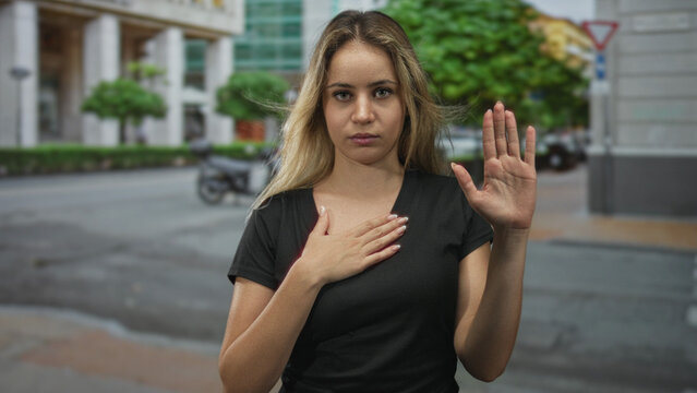 Young blonde woman wearing black t shirt placing hand on chest with palm raised in oath gesture while standing on busy city street near buildings; pledge serenity.