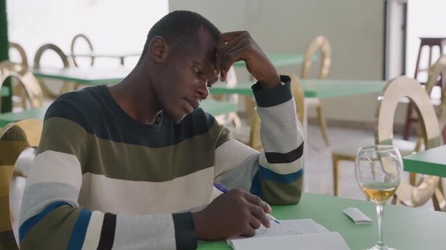 Black man writing after date note with wine glass on table, restaurant bistro setting with empty chairs, melancholic expression as he drafts apology and rereads memories, late evening mood of regret