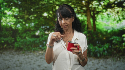 Obraz premium Senior hispanic woman in white blouse holds a red coffee mug with her right hand, puckers lips and glances aside while standing among leafy trees in a forest; contemplation.