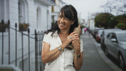 Fototapeta premium Woman holding pottery bottle to chest with hands on street sidewalk by rowhouse railing, smiling as she holds the vase; contentment.