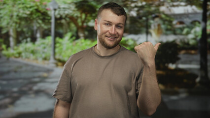 Fototapeta premium Man pointing thumb to the side with bare hand visible in a forest park setting, smiling and relaxed; friendly casual.
