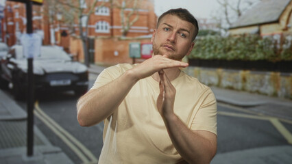 Fototapeta premium Man making timeout gesture with hands on a city street in front of brick building and parked car; frustration warning.