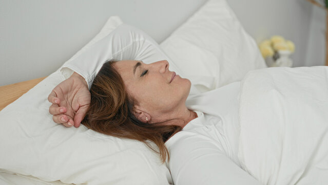 Woman lying supine in bed with arm over forehead, hand near temple, eyes closed, pillow and white duvet, bedside plant and vase visible in bedroom building; calm rest.