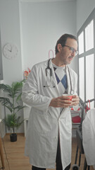 Fototapeta premium Man doctor in white coat holds red coffee cup with both hands and stethoscope at neck in medical building; quiet concern.