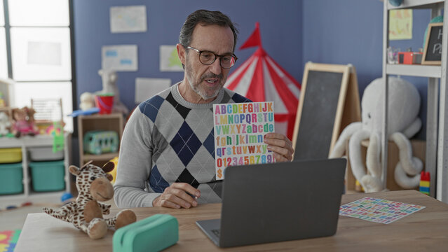 Man holding colorful alphabet chart and pointing with pen at letters beside laptop in school building; teaching focus.