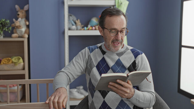 Man reads book with hand on crib in baby room studio, holding pages and touching railing; quiet bedtime serenity.