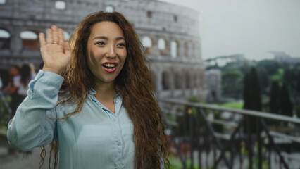 Obraz premium Smiling woman waving her hand at the coliseum building on an outdoor terrace railing near ancient arches and trees; travel joy.