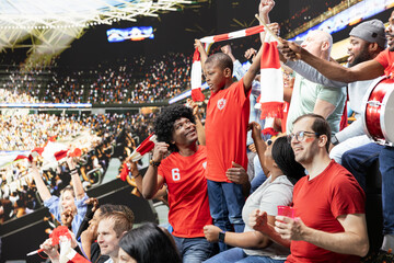 Fototapeta premium Excited parents and their little boy enjoy live football match together, surrounded by stadium crowd energy and passionate supporters. Black family sharing family outing with cheering.