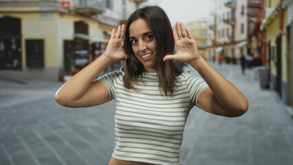 Fototapeta premium Woman frames face with hands while smiling on a busy pedestrian street lined with storefronts and cobblestone pavement, wearing a striped t shirt and casual style; playful energy.