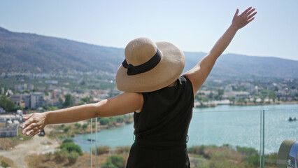 Obraz premium Woman with outstretched arms and straw sunhat on building deck overlooking turquoise sea and coastal town, facing away from camera; serenity escape.