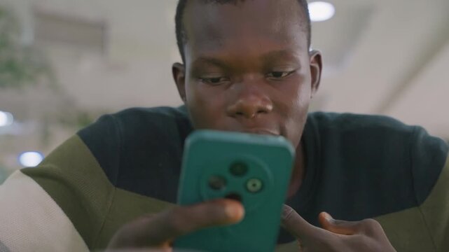 Black man checking phone closely, thumbs on screen and lip bite, teal phone case visible, concentrated expression under warm indoor lights, intimate closeup capturing anxiety and hopeful anticipation