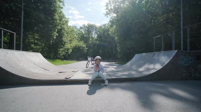 Little girl in a safety helmet sitting on a skate ramp and drinking water from a bottle after riding a bicycle