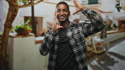 Fototapeta premium Young black man smiling and framing face with hands in a plaid shirt on a sunlit street; playful confidence.