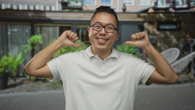 Young man wearing glasses and a white polo pointing thumbs to chest at a street entrance of a house; self confidence.