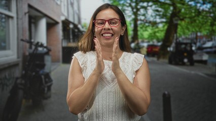 Fototapeta premium Woman clapping hands and smiling on city street with red glasses and white top; joy gratitude warmth contentment.