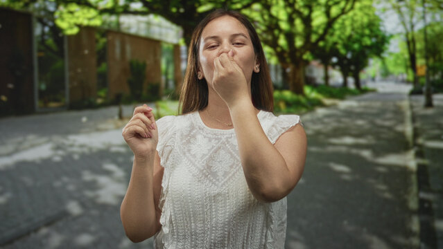 Woman pinches nose with fingers on a tree lined street beside a building entrance in daylight; disgust reaction.