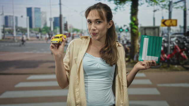 Young woman smiling, holding a yellow toy car and a learner plate at a city street crosswalk; learning independence confidence.